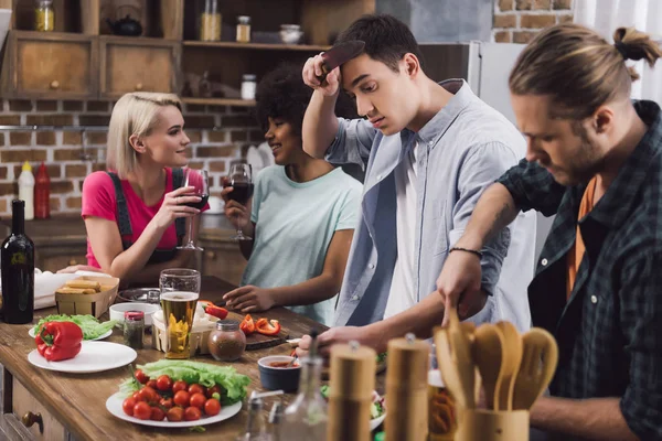 Filles multiethniques boire du vin tandis que les hommes cuisine dans la cuisine — Photo de stock