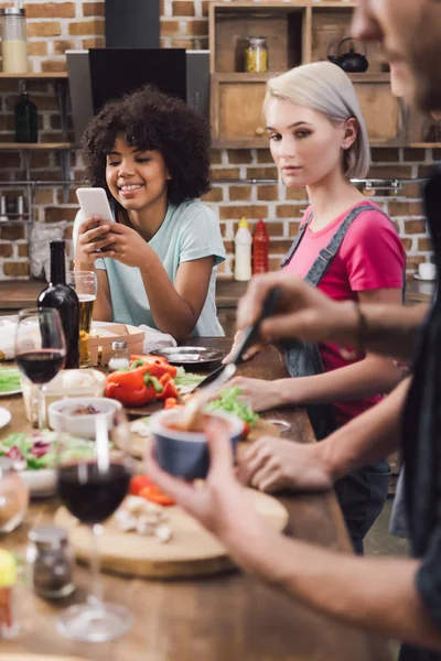 Smiling african american girl looking at smartphone while friends cooking — Stock Photo