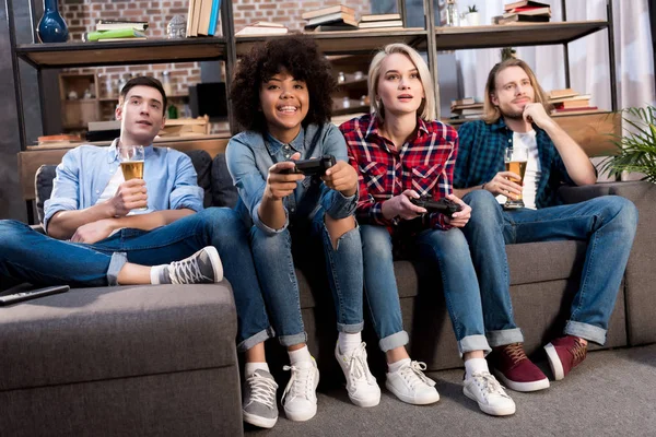 Мультикультурные девушки играют в видеоигры на диване дома, мужчины пьют пиво — стоковое фото