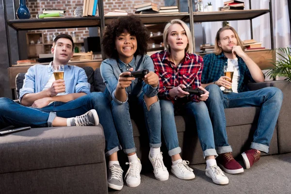 Multicultural girls playing video game on sofa at home, men drinking beer — Stock Photo