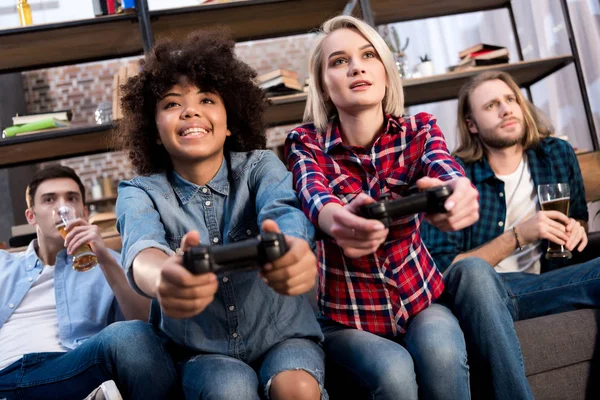 Multicultural girls playing video game on sofa at home, men drinking beer — Stock Photo