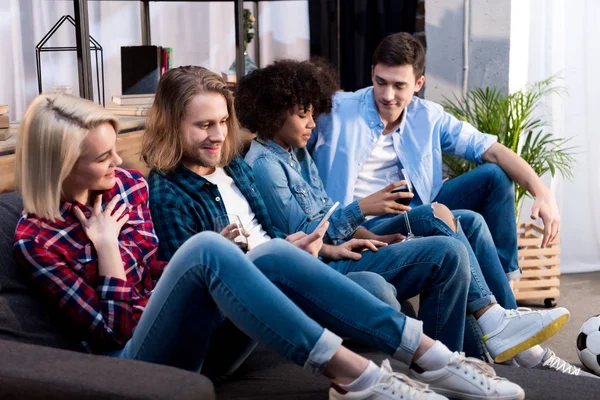 Multicultural friends sitting on sofa and looking at smartphone — Stock Photo