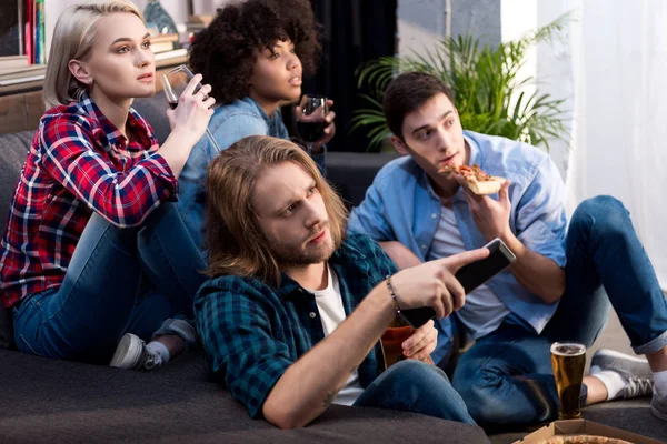 Amis multiculturels regarder la télévision avec de l'alcool et de la nourriture à la maison — Photo de stock