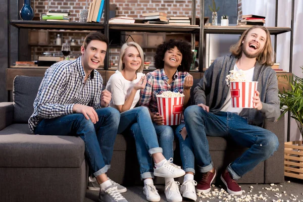 Laughing multicultural friends watching tv on sofa with popcorn — Stock Photo