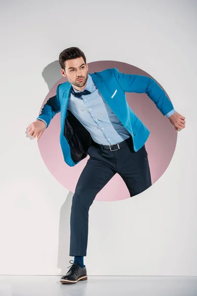 Guapo joven con estilo en traje y corbata de lazo mirando hacia otro lado mientras camina a través del agujero en gris - foto de stock