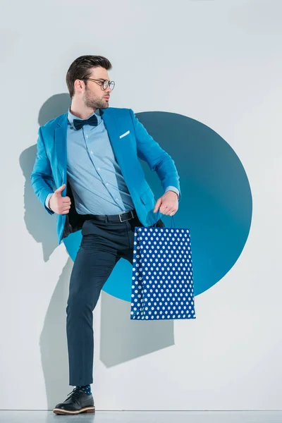 Guapo joven con estilo con bolsa de compras mirando hacia otro lado mientras se mete a través del agujero en gris - foto de stock