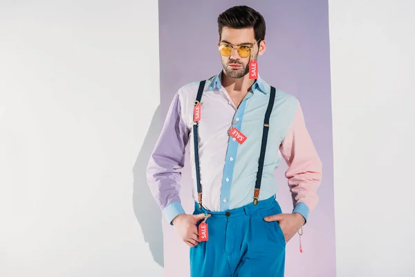 Beau jeune homme élégant en lunettes de soleil avec des étiquettes de vente sur gris — Photo de stock