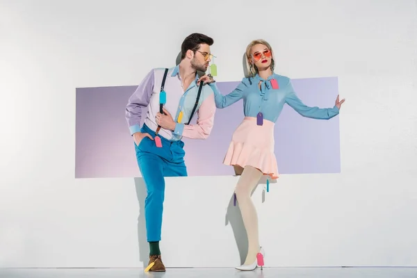 Fashionable young couple with colorful tags on clothes walking through opening on grey — Stock Photo