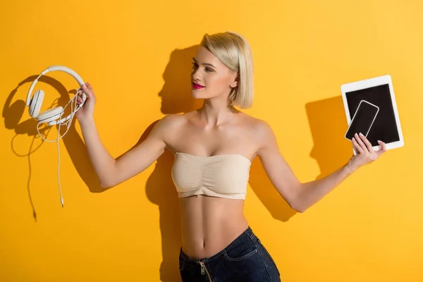 Schöne lächelnde blonde Mädchen mit Kopfhörer, Smartphone und digitalem Tablet auf gelb — Stockfoto