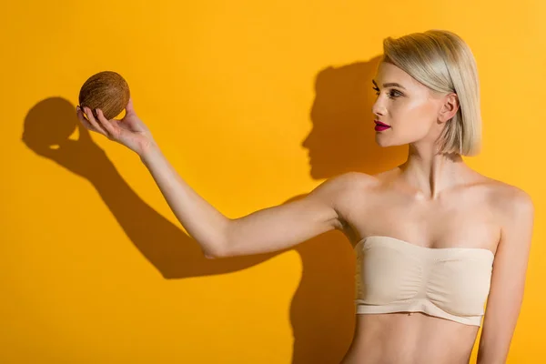 Красива молода блондинка вгорі тримає кокос на жовтому — стокове фото