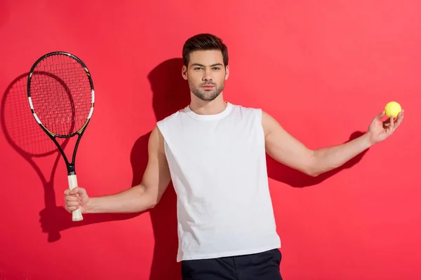 Jovem bonito segurando raquete de tênis com bola e olhando para a câmera em rosa — Fotografia de Stock