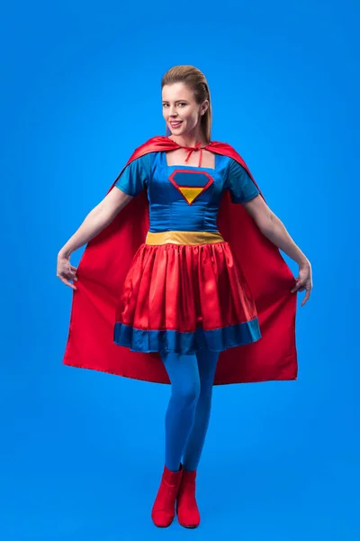 Mulher atraente em traje de super-herói segurando capa isolada em azul — Fotografia de Stock