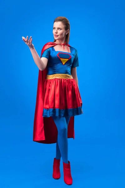 Atractiva mujer en traje de superhéroe con gestos de capa aislado en azul - foto de stock