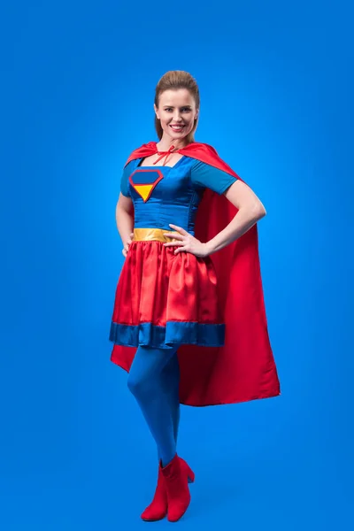 Bela mulher sorridente em traje de super-herói de pé akimbo isolado em azul — Fotografia de Stock