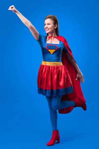 Mulher sorridente em traje de super-herói com braço estendido isolado em azul — Fotografia de Stock