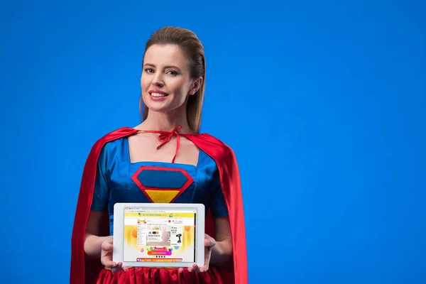 Retrato de mulher sorridente em traje de super-herói mostrando tablet isolado em azul — Fotografia de Stock
