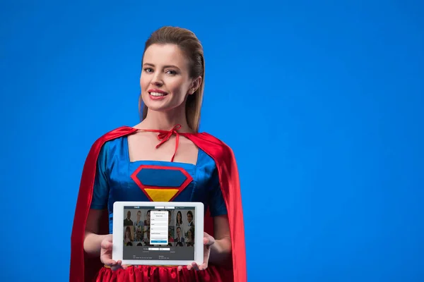 Portrait of cheerful woman in superhero costume showing tablet isolated on blue — Stock Photo