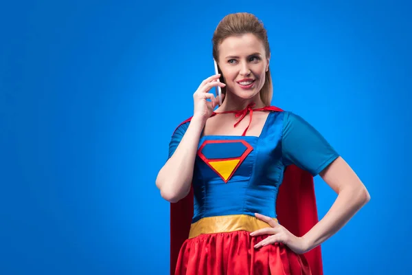 Portrait of smiling woman in superhero costume talking on smartphone isolated on blue — Stock Photo