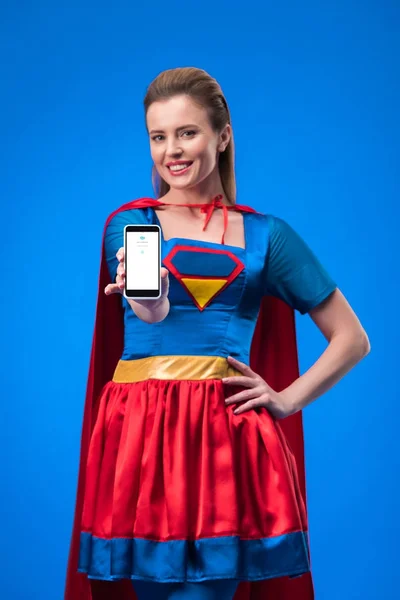 Portrait of smiling woman in superhero costume showing smartphone isolated on blue — Stock Photo