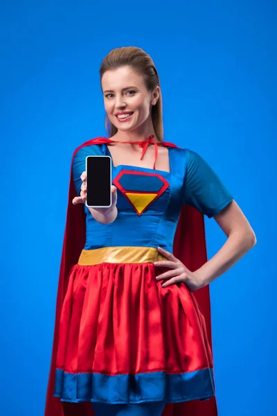 Ritratto di donna sorridente in costume da supereroe che mostra smartphone con schermo bianco isolato su blu — Foto stock