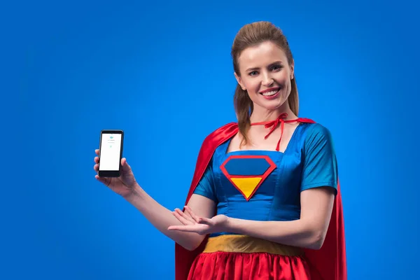 Portrait of smiling woman in superhero costume showing smartphone isolated on blue — Stock Photo
