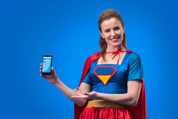 Ritratto di donna sorridente in costume da supereroe che mostra smartphone isolato su blu — Foto stock