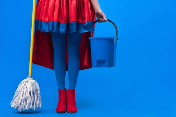 Vista parcial de la mujer en traje de superhéroe con fregona y cubo para la limpieza aislado en azul - foto de stock