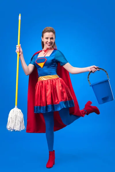 Mujer feliz en traje de superhéroe con fregona y cubo para limpiar aislado en azul - foto de stock