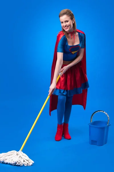 Cheerful woman in superhero costume with mop and bucket for cleaning isolated on blue — Stock Photo