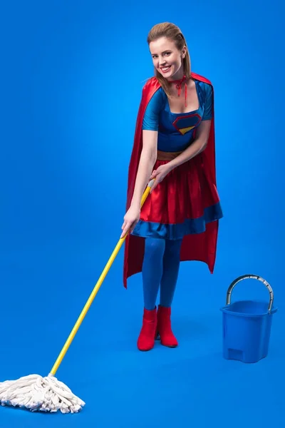 Mulher alegre em traje de super-herói com esfregona e balde para limpeza isolada em azul — Fotografia de Stock