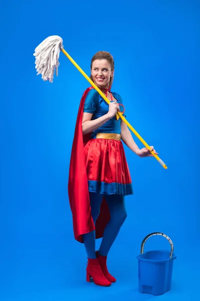 Mujer sonriente en traje de superhéroe con fregona y cubo para limpiar aislado en azul - foto de stock