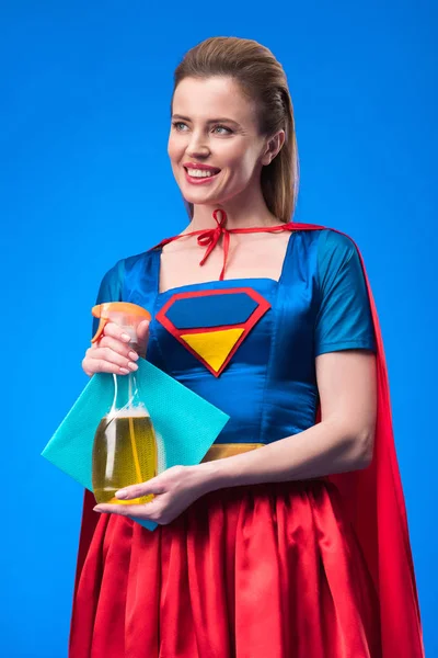 Portrait of smiling woman in superhero costume with rag and detergent in hands isolated on blue — Stock Photo