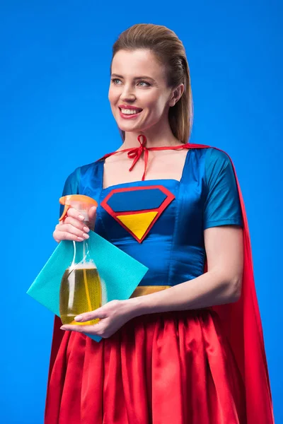 Портрет улыбающейся женщины в костюме супергероя с тряпкой и моющим средством в руках, изолированных на голубом — стоковое фото