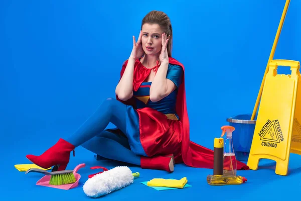 Mulher cansada em traje de super-herói com suprimentos de limpeza em torno isolado em azul — Fotografia de Stock
