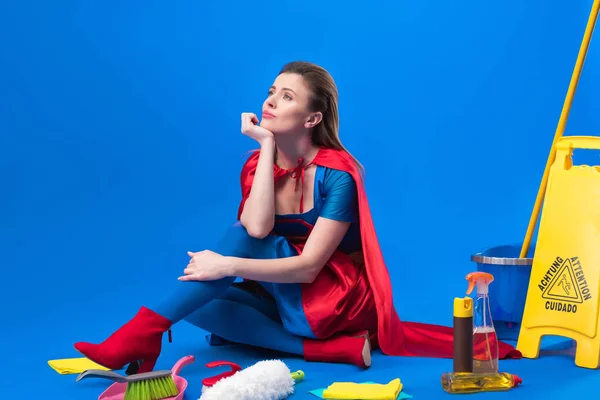 Femme coûteuse en costume de super-héros avec des fournitures de nettoyage autour isolé sur bleu — Photo de stock