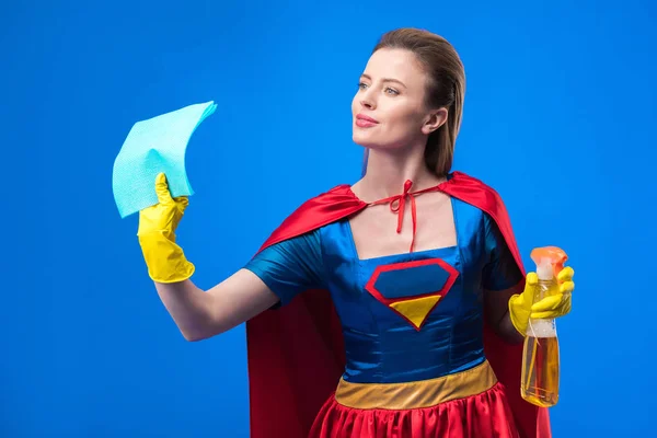 Portrait of woman in superhero costume and rubber gloves with rag and detergent isolated on blue — Stock Photo