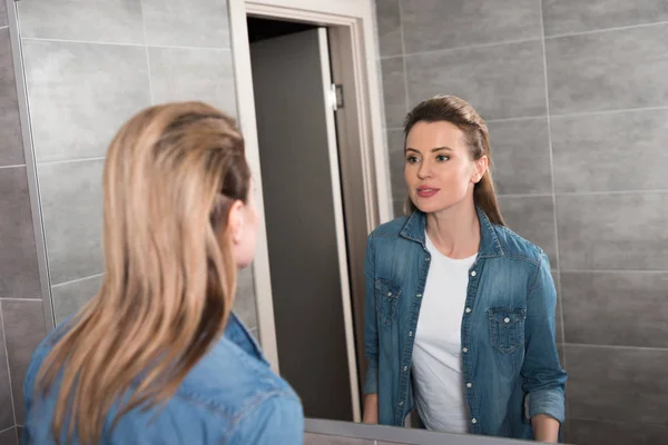 Attraente donna in abbigliamento casual guardando nello specchio a casa — Foto stock