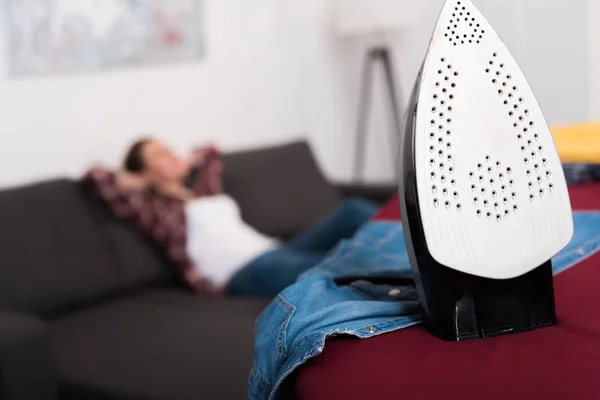 Selective focus of iron on ironing board and woman resting on sofa — Stock Photo