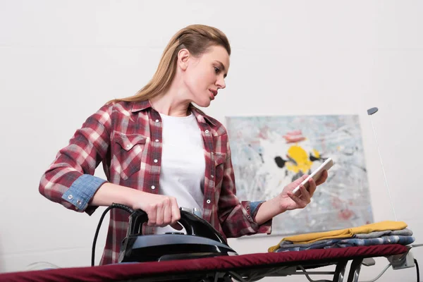 Donna utilizzando smartphone mentre stirare i vestiti a casa — Foto stock