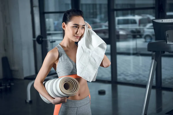 Junge sportliche Frau mit gerollter Yogamatte wischt nach Übungen im Fitnessstudio Schweiß mit Handtuch ab — Stockfoto