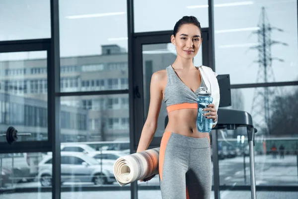 Jeune femme sportive avec tapis de yoga roulé et l'eau à la salle de gym — Photo de stock