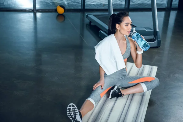 Exhausted athletic woman drinking water while sitting on yoga mat at gym — Stock Photo