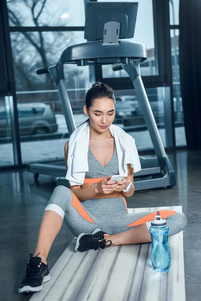 Young sporty woman using smartphone while sitting on yoga mat at gym — Stock Photo