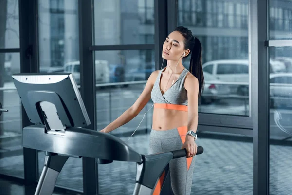 Young athletic sportswoman jogging on treadmill and listening music with earphones at gym — Stock Photo