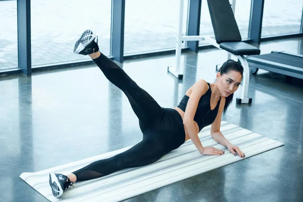 Joven mujer deportiva levantando pierna en esterilla de yoga en el gimnasio - foto de stock
