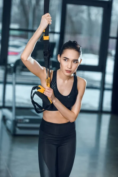 Jovem mulher desportiva exercitando-se relaxante no ginásio, enquanto se inclina em tiras de suspensão — Fotografia de Stock