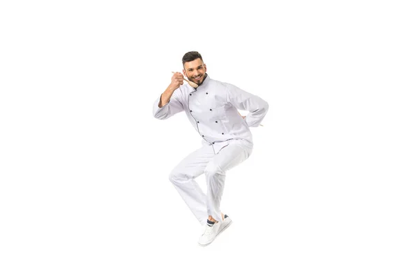 Young male chef tasting food with spoon while jumping and smiling at camera isolated on white — Stock Photo