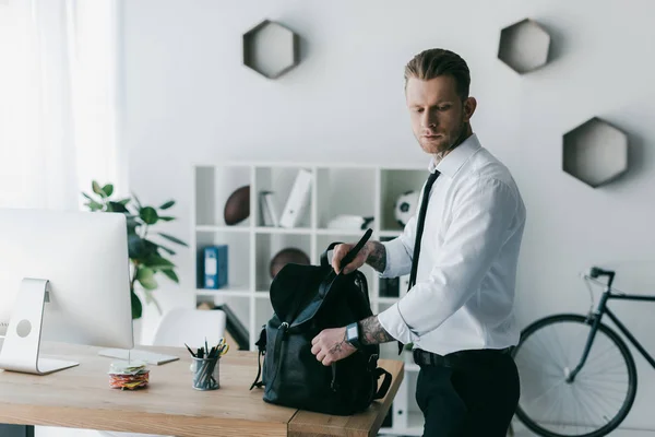 Schöner junger tätowierter Geschäftsmann mit Rucksack, der im Büro nach unten schaut — Stockfoto