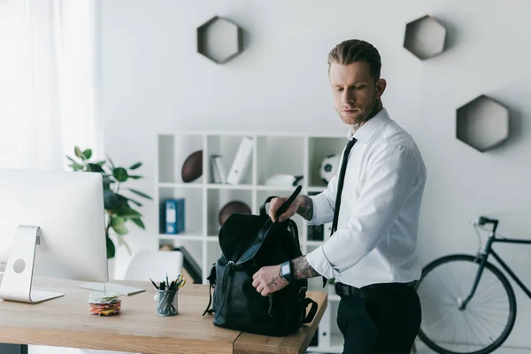 Beau jeune homme d'affaires tatoué avec sac à dos regardant en bas dans le bureau — Photo de stock