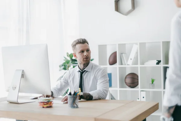 Guapo joven hombre de negocios tatuado mirando hacia otro lado mientras trabaja con la computadora de escritorio en la oficina - foto de stock